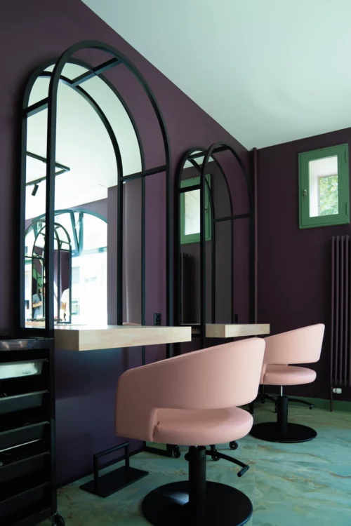 Poste de coiffure chez Euphoria Hair & Beauty Studio à Carouge (Genève) : fauteuils rose poudré, miroirs à arches en métal noir, murs prune.