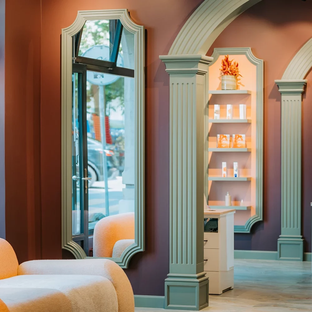 Intérieur d’Euphoria Hair & Beauty Studio à Carouge : arches et moulures vert sauge, grand miroir, présentoir de produits, banquette claire près de la vitrine.