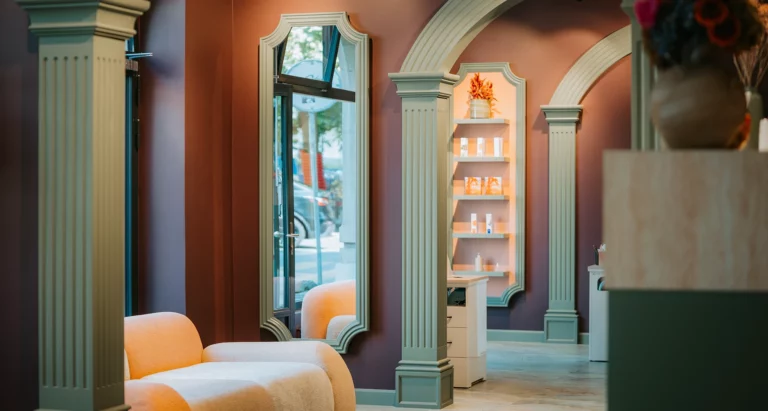 Intérieur d’Euphoria Hair & Beauty Studio à Carouge : arches et moulures vert sauge, grand miroir, présentoir de produits, banquette claire près de la vitrine.