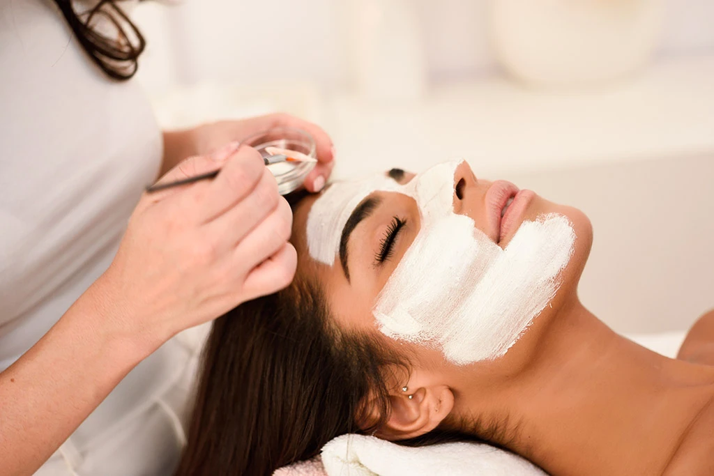 Application d’un masque visage blanc au pinceau sur une femme allongée lors d’un soin en institut.